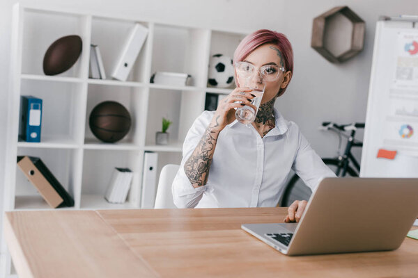 young tattooed businesswoman in eyeglasses drinking water while using laptop in office