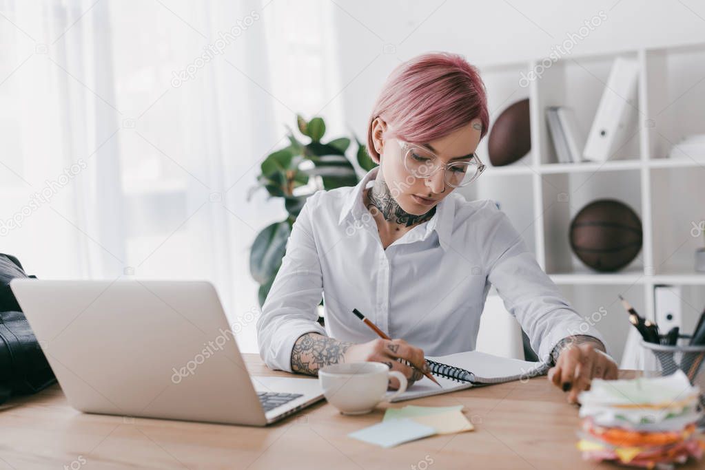 Young tattooed businesswoman taking notes while using laptop at workplace