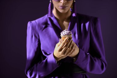 Cropped view of african american girl in ultra violet jacket with cupcake, isolated on dark purple clipart