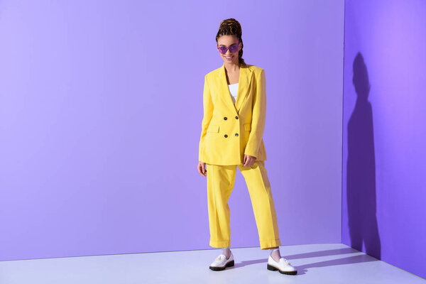 улыбающаяся африканская американская девушка в жёлтом костюме и фиолетовых солнцезащитных очках, на модном ультрафиолетовом фоне
