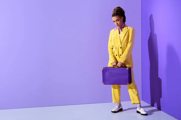 紫のスーツケースとトレンディな黄色いスーツでポーズをとってアフリカ系アメリカ人の女の子 — ストック写真