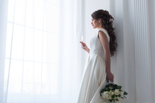 brunette bride in dress with wedding bouquet and glass of champagne looking at window