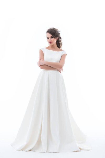 привлекательная невеста позирует в традиционном свадебном платье со скрещенными руками, изолированная на белом
