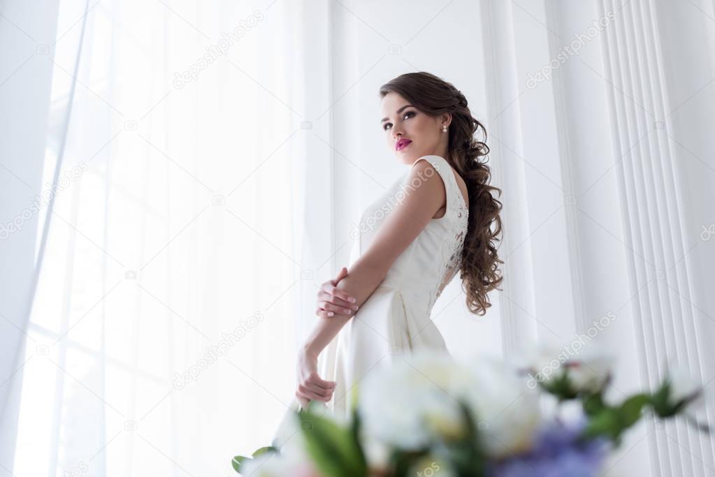 Brunette caucasian bride in wedding dress at window, flowers on foreground