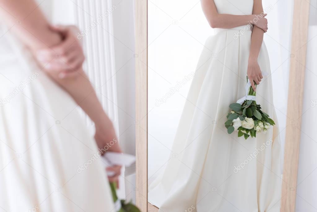 cropped view of bride in traditional dress with wedding bouquet looking at her reflection in mirror