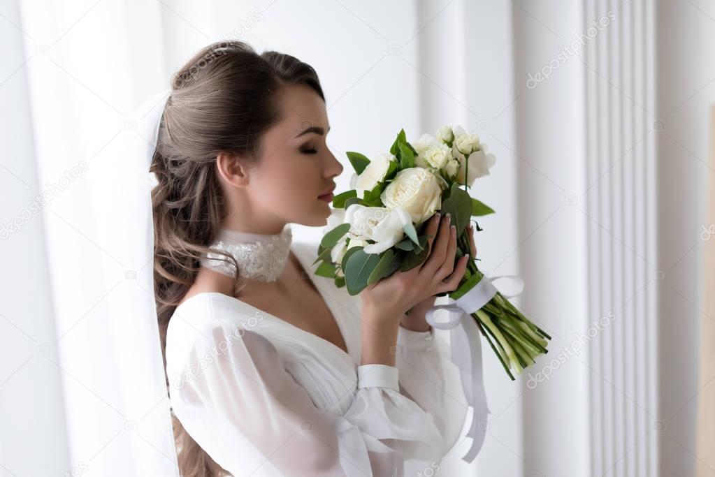 attractive bride in wedding dress and veil sniffing white bouquet
