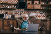 Entspannter alter Mann mit Kopfhörer und Laptop mit Füßen auf dem Tisch