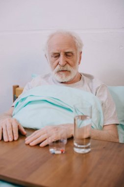 Old man looking sadly at pills on table clipart