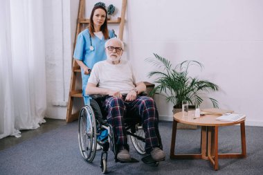 Tekerlekli sandalyede üst düzey erkeğin arkasında duran kadın hemşire