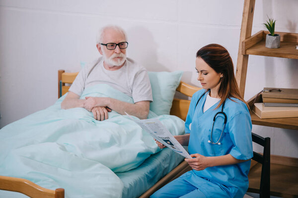 Old man patient looking at nurse reading newspaper
