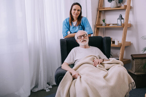 Улыбающаяся медсестра, стоящая позади пожилого мужчины в кресле
