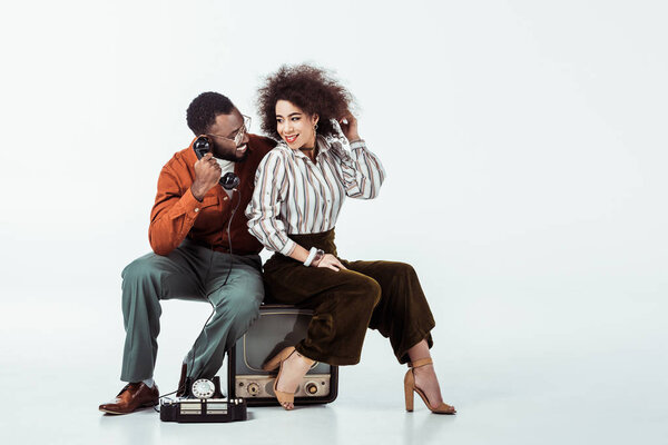 happy african american retro styled couple sitting on vintage television with phone on white