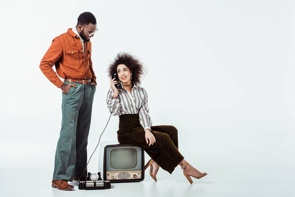 african american retro styled girlfriend sitting on vintage television and and talking by phone on white