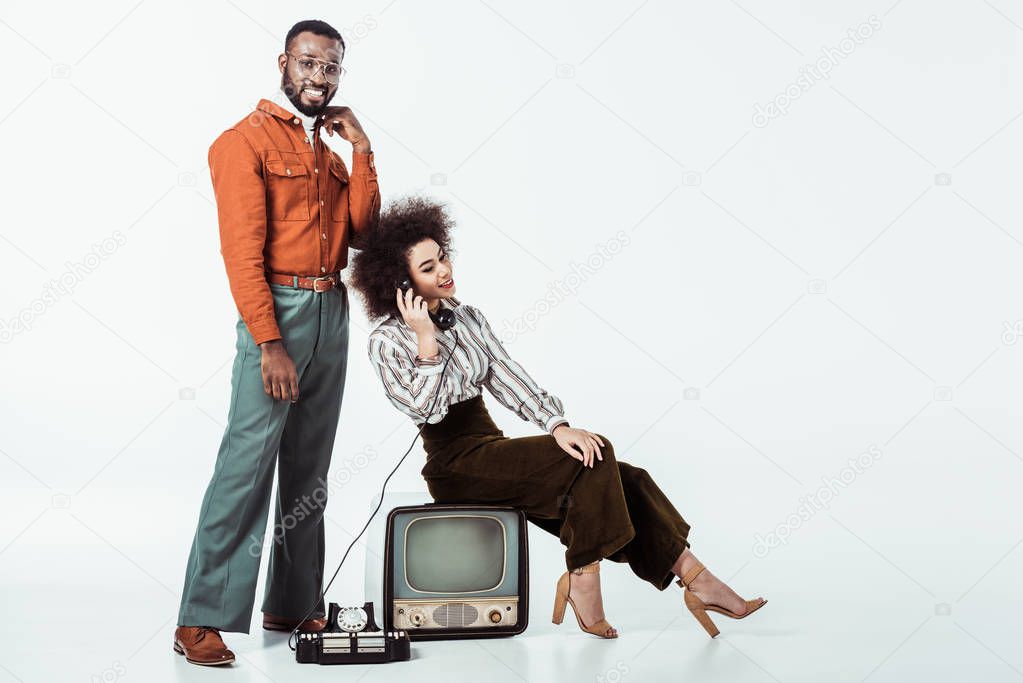 happy african american retro styled girlfriend sitting on vintage television and and talking by phone on white