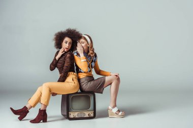 multicultural retro styled girls gossiping and sitting on vintage television clipart