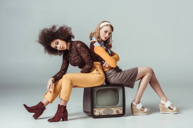 multicultural retro styled girls sitting on vintage television and looking at camera on grey clipart