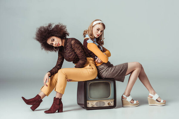 multicultural retro styled girls sitting on vintage television and looking at camera on grey