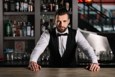 handsome bartender leaning on bar counter and looking at camera clipart