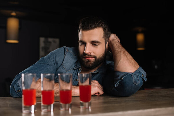 handsome visitor leaning on bar counter and looking at four shot drinks