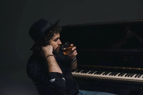 Guapo músico bebiendo whisky cerca de piano en negro —  Fotos de Stock