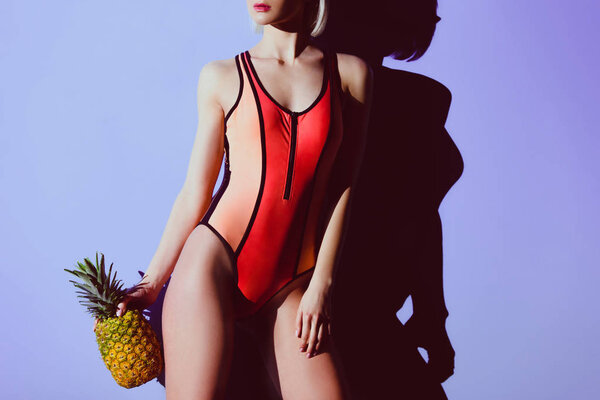 cropped view of slim girl posing in swimsuit with pineapple, on purple