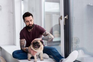 tattooed handsome man sitting on windowsill and palming dog at home clipart