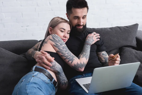 happy tattooed couple looking at laptop on sofa at home