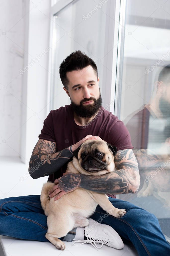 tattooed handsome man sitting on windowsill and hugging pug at home
