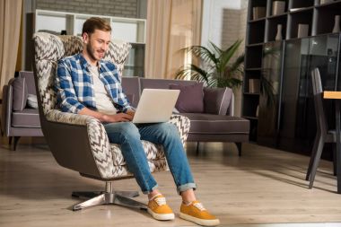 Young male in armchair using laptop in living room with modern design clipart