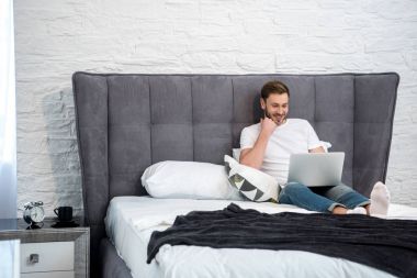 Happy young male using laptop in cozy modern bedroom clipart