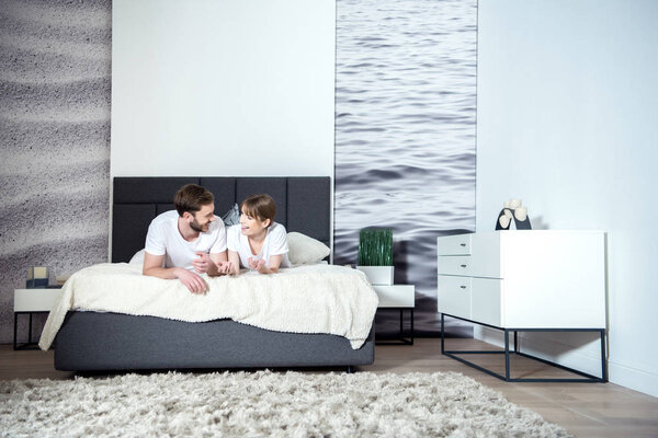 Smiling couple talking and lying on bed in cozy bedroom with modern interior