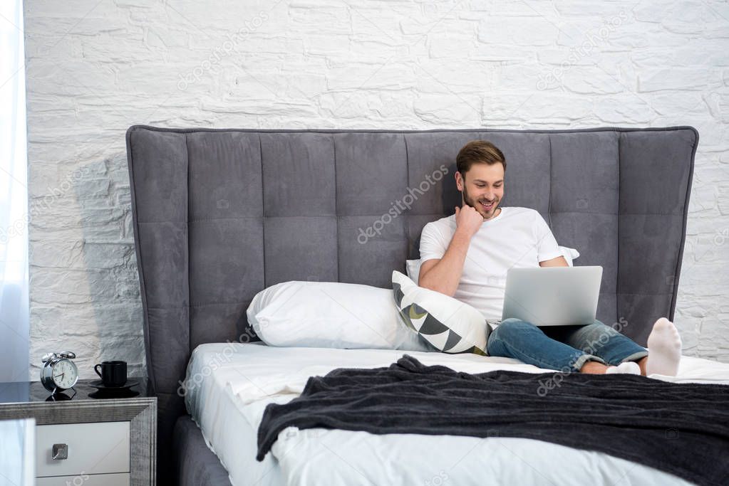 Happy young male using laptop in cozy modern bedroom