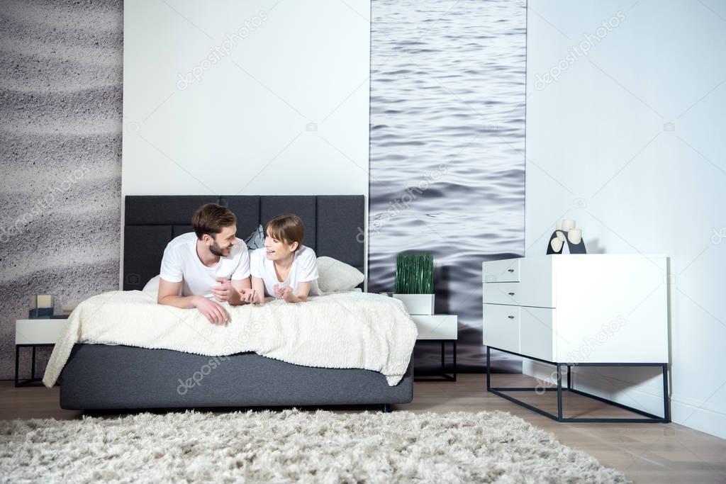 Smiling couple talking and lying on bed in cozy bedroom with modern interior