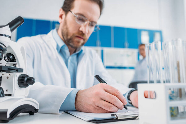 selective focus of scientist in white coat and eyeglasses making notes in notepad at workplace in laboratory