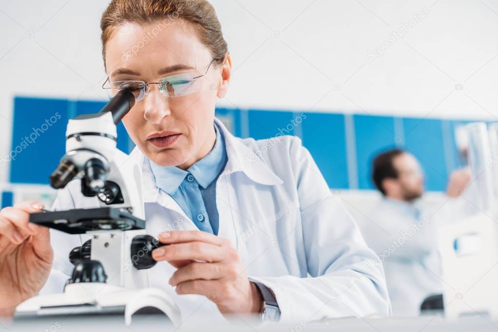 selective focus of female scientist looking thorough microscope on reagent with colleague behind in lab
