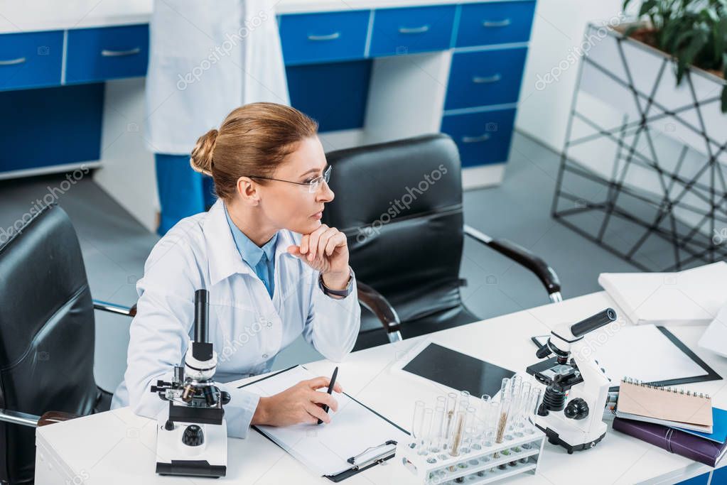 selective focus of pensive female researcher at workplace with colleague behind in lab