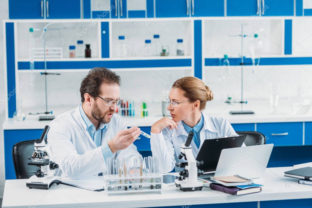 portrait of scientific researchers in white coats at workplace with flasks, microscopes and laptop in laboratory