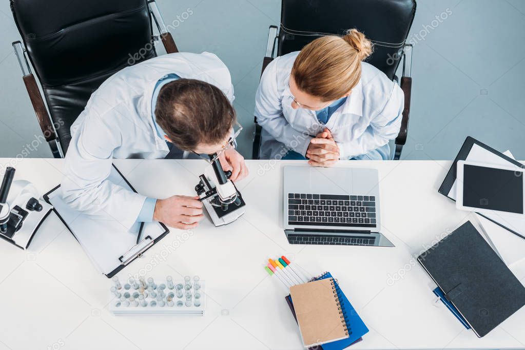 overhead view of scientific researchers in white coats working together at workplace with microscope and laptop in laboratory