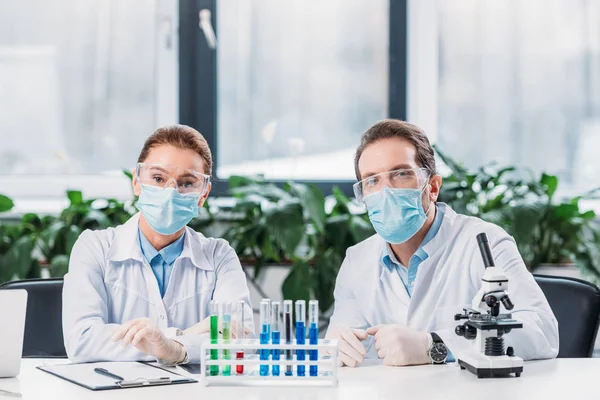 scientific researchers in goggles and medical masks sitting at workplace with reagents in tubes and microscope in lab