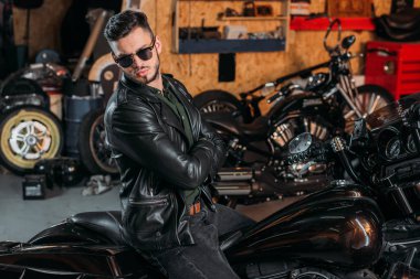 handsome young man in sunglasses and leather jacket on bike at garage clipart