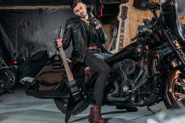 stylish young man in leather jacket sitting on bike with electric guitar at garage clipart