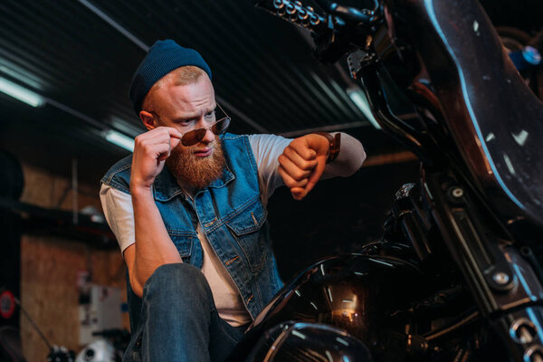 handsome young man in sunglasses looking at watch while sitting on motorcycle at garage