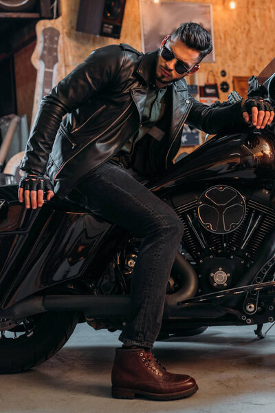 handsome young man in stylish clothing sitting on motorcycle at garage