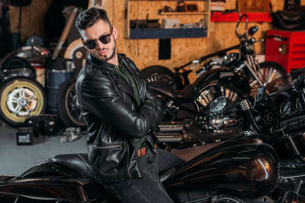 handsome young man in sunglasses and leather jacket on bike at garage