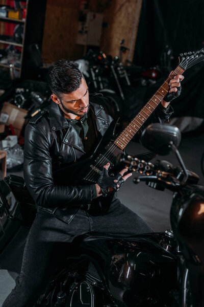 handsome young man playing guitar while sitting on bike at garage