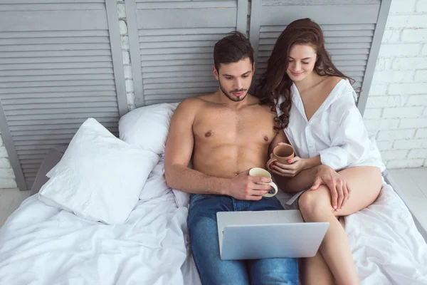 Jovem Casal Com Xícaras Café Usando Laptop Enquanto Descansa Cama — Fotografia de Stock