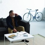 Bearded senior man talking by smartphone while sitting on sofa and looking away at home