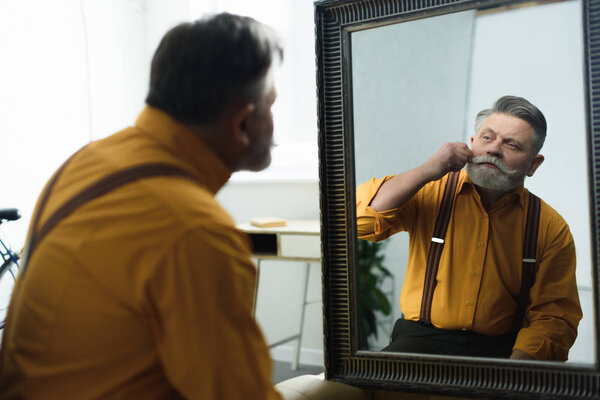 handsome bearded senior man adjusting moustache and looking at mirror