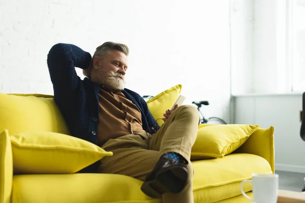 スマート フォンを使用して自宅のソファーに座っている年配の男性にひげを生やした笑顔 — ストック写真