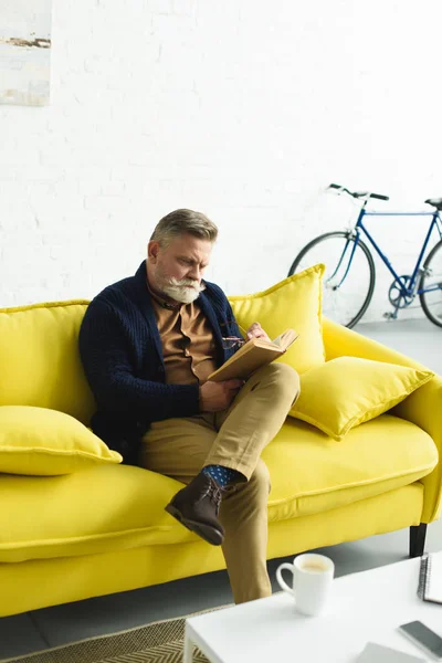 Γενειοφόρος Ανώτερος Άνθρωπος Κάθεται Στο Κίτρινο Καναπέ Και Διαβάζοντας Βιβλίο — Δωρεάν Φωτογραφία